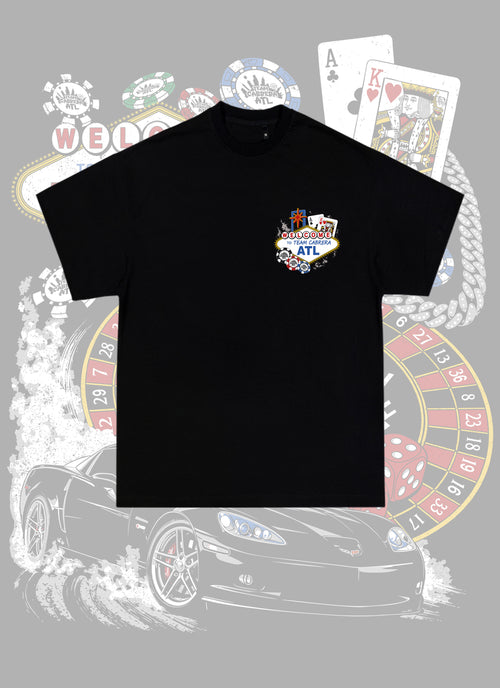 A black short-sleeve t-shirt with a colorful 'Welcome to ATL' casino-themed graphic on the left chest, displayed over a gray background with a faded illustration of a sports car, roulette wheel, and playing cards.
